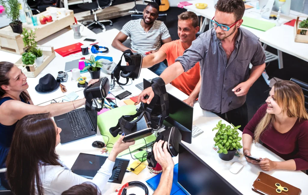 Team of coworkers at a desk laughing while passing around a VR headset among laptops and office supplies
